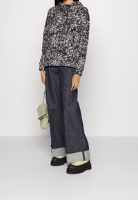 Blusa navy con estampado floral y capucha, combinada con pantalones de mezclilla oscuros y holgados con dobladillos enrollados. Zapatos de plataforma negros y un bolso ligero verde.