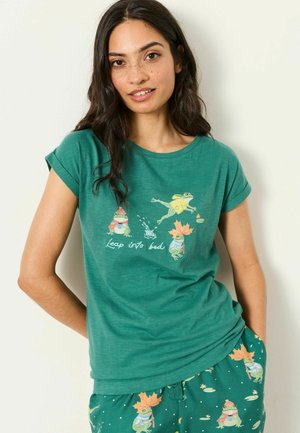 Woman with long dark hair wearing green pajamas with cartoon frogs and the text "Leap into bed" on the shirt.