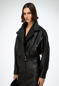 Mujer con cabello corto y castaño que lleva puesto un abrigo de cuero negro de gran tamaño y una falda a juego, sosteniendo un bolso negro de piel sintética.