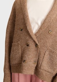 Bruine gebreide cardigan met een V-hals, dubbele knoopsluiting, twee frontzakken en grote ronde knopen. Gelaagd over een roze kledingstuk.