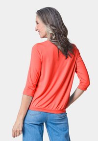 Korallfarbener, leichter Oberteil mit 3/4-Ärmeln und gerafftem Saum. Verfügt über eine glatte Textur und ein lässiges Design. Kombiniert mit blauen Jeans.