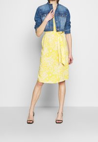 Gul blommig klänning med knytning i midjan, kombinerad med en åtsittande jeansjacka. Klänningen har vita mönster; jackan har två bröstfickor.