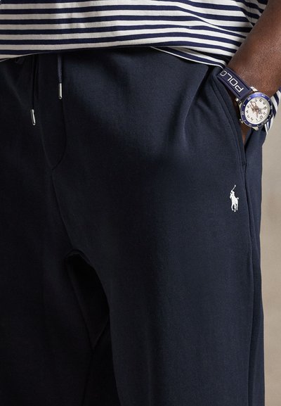 Person iført marineblå joggingbukser med hvidt Polo-logo og stribet skjorte, hånd i lommen, iført blå Polo-armbåndsur.