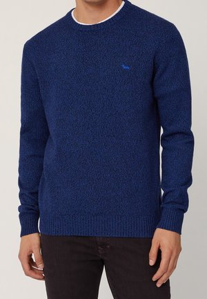 Mężczyzna ubrany w granatowy sweter o fakturowanej powierzchni z prążkowanymi mankietami i dolnym ściągaczem, w połączeniu z ciemnymi spodniami.