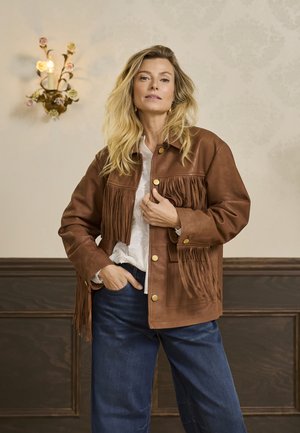 Brun læderjakke med frynse detaljer, gyldne knapper og en afslappet pasform, båret over en hvid bluse med et tekstureret mønster. Blå denim bukser.