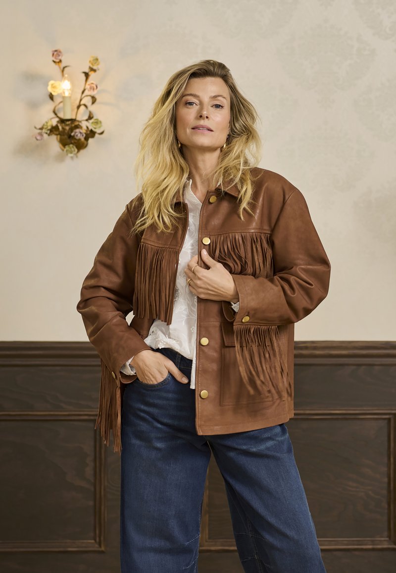 Brun læderjakke med frynse detaljer, gyldne knapper og en afslappet pasform, båret over en hvid bluse med et tekstureret mønster. Blå denim bukser.