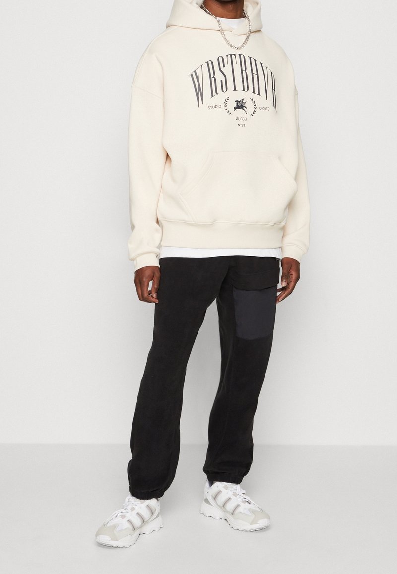 Felpa crema con stampa del logo, tasca anteriore, abbinata a pantaloni jogger neri con tasca laterale, indossata con sneakers bianche.
