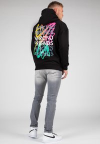 Felpa nera con il testo colorato "WRONG FRIENDS" sul retro. Il tessuto sembra morbido e il design presenta lettere sovrapposte e vivaci. Abbinata a jeans grigi chiari e sneaker bianche.