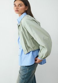 Jasnozielona krótka kurtka z elastycznymi mankietami, noszona na niebieskiej i białej w paski koszuli zapinanej na guziki, w połączeniu ze średnio zniszczonymi dżinsami.