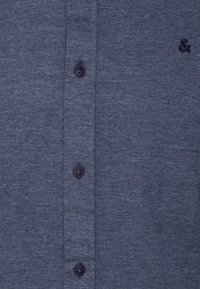 Mörkblå skjorta med knappar i strukturerad tyg, som har sex svarta knappar och en liten broderad logotyp nära kragen.