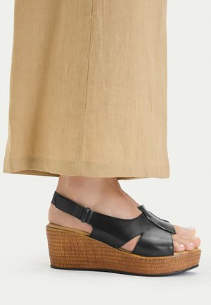 Weiblicher Fuß mit schwarzen Leder-Slingback-Sandalen mit offenem Zehenbereich, Keilabsatz und holzstrukturierter Sohle sowie beigen Leinen-Hosen mit weitem Bein.