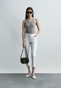 Kvinne iført svarte solbriller, grå ermeløs topp, hvite høytlivsbukser med knapper, hvite hæler, holder svart veske, stående mot lys bakgrunn.