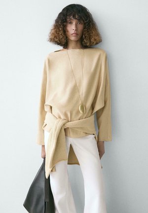 Femme aux cheveux bouclés portant un haut beige, un pull noué autour de la taille, un pantalon blanc, un long pendentif en or, tenant un sac noir.