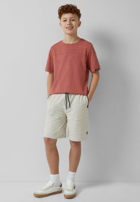 Helles rosa T-Shirt mit kurzen Ärmeln und Nähdetails, kombiniert mit cremefarbenen Shorts mit elastischem Bund und schwarzem Kordelzug sowie weißen Turnschuhen.