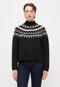Zwarte gebreide trui met een hoge hals, voorzien van een wit geometrisch patroon rond de schouders. Geribbelde manchetten en zoom.