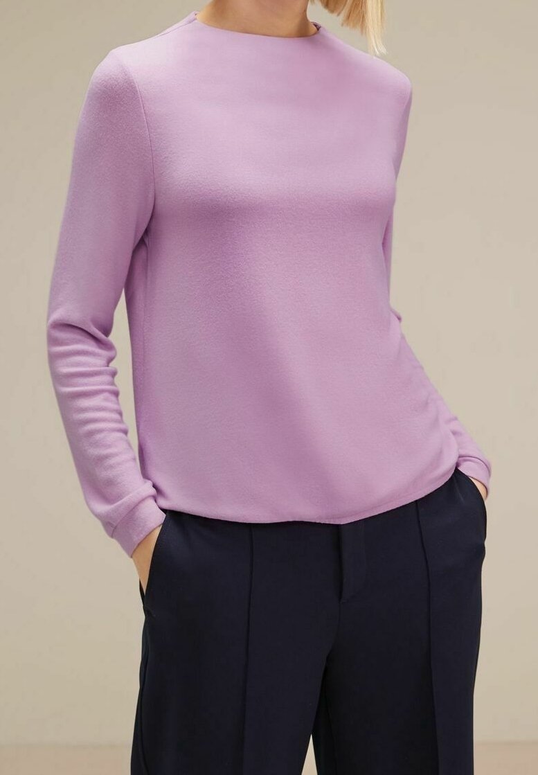 Femme portant un pull lavande à manches longues rentré dans un pantalon noir taille haute, les mains dans les poches, sur un fond neutre.