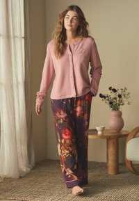 Roze knoop-hals cardigan met lange mouwen, gecombineerd met luchtige, bloemenpatroon pyjamabroeken in paarse en oranje tinten. Zachte texturen zichtbaar.
