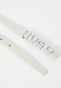Cintura in pelle bianca con un prominente logo "HUGO" in argento, caratterizzata da una superficie liscia e fori regolabili per la vestibilità.