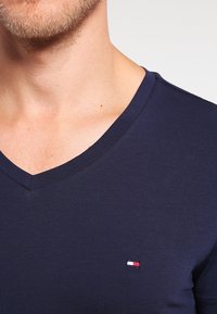 Camiseta de algodón azul marino con cuello en V y forma ajustada. Presenta un pequeño logo bordado en rojo, blanco y azul en el lado izquierdo del pecho.
