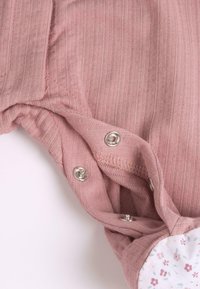 Body en rose côtelé avec des boutons-pression argentés en bas. Présente une doublure blanche à motif floral. Texture douce adaptée au confort.