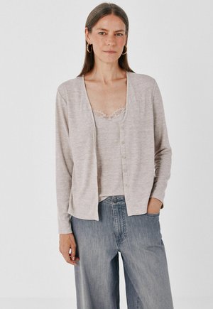 Femme portant un cardigan beige boutonné sur un caraco orné de dentelle et un jean bleu ample, debout avec une main dans la poche sur fond blanc.