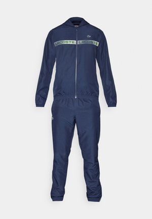 Ensemble de survêtement bleu marine en tissu lisse, comprenant une veste zippée avec capuche et un pantalon assorti. Détails de logo verts sur la poitrine.