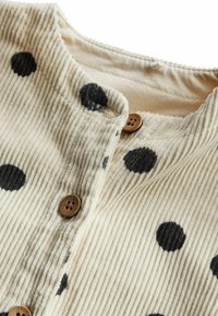 Beige corduroy shirt met zwarte stippen, voorzien van een ronde kraag en twee bruine knoopsluitingen. Gestructureerde verticale strepen.