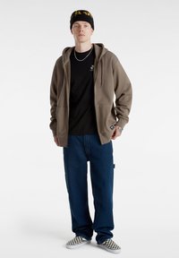 Sudadera con cremallera marrón sobre una camiseta negra, jeans holgados azules y zapatillas de cuadros en blanco y negro; presenta un gorro negro con detalles amarillos.