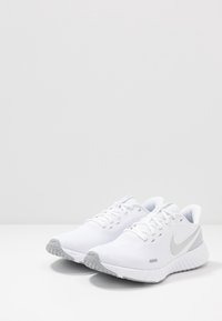 Chaussures de sport blanches avec une tige en mesh texturé, des accents gris et une semelle rembourrée. Présentent un logo bien visible et un système de laçage traditionnel.