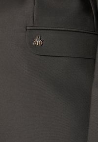 Tmavě šedý sako s elegantním designem, hladkým povrchem a malým kovovým logem na kapse. Přiléhavý střih s čistými liniemi.