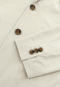 Camisa de un beige claro con un diseño elegante, que cuenta con cuatro botones oscuros y estampados en el puño y un cierre frontal de dos botones.