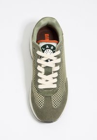 Sneaker verde oliva con materiali in rete e suede, lacci crema, patch circolare con logo e una soletta marrone. Presenta una suola testurizzata.