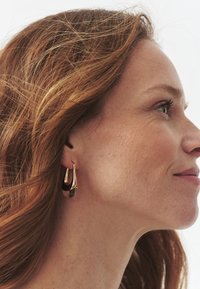 Ohrhänger in Goldoptik mit einem glatten schwarzen Akzent, gestaltet mit fließenden Kurven und einem Hakenverschluss, am Ohr des Modells präsentiert.