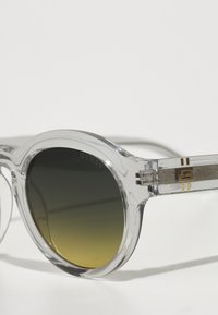 Ochelari de soare din plastic transparent cu lentile rotunde, nuanță gradient de verde la galben, accente aurii pe brațe și un logo gravat pe o lentilă.