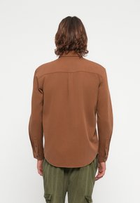 Bruine lange mouwen shirt met een gebogen onderrand, knoopmanchetten en een schouderpas aan de achterkant. Het materiaal lijkt zacht met een gladde textuur.