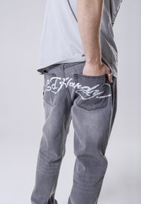 Šedé džíny z denimu s opotřebovaným vzhledem a bílým vyšitým textem na zadních kapsách. Design zahrnuje slim fit a standardní kapsy.