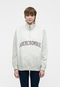 Młoda osoba z krótkimi brązowymi włosami, nosząca jasnoszary sweatshirt z ćwierć suwakowym zamkiem i napisem "ABERCROMBIE" oraz ciemnoniebieskie dżinsy.