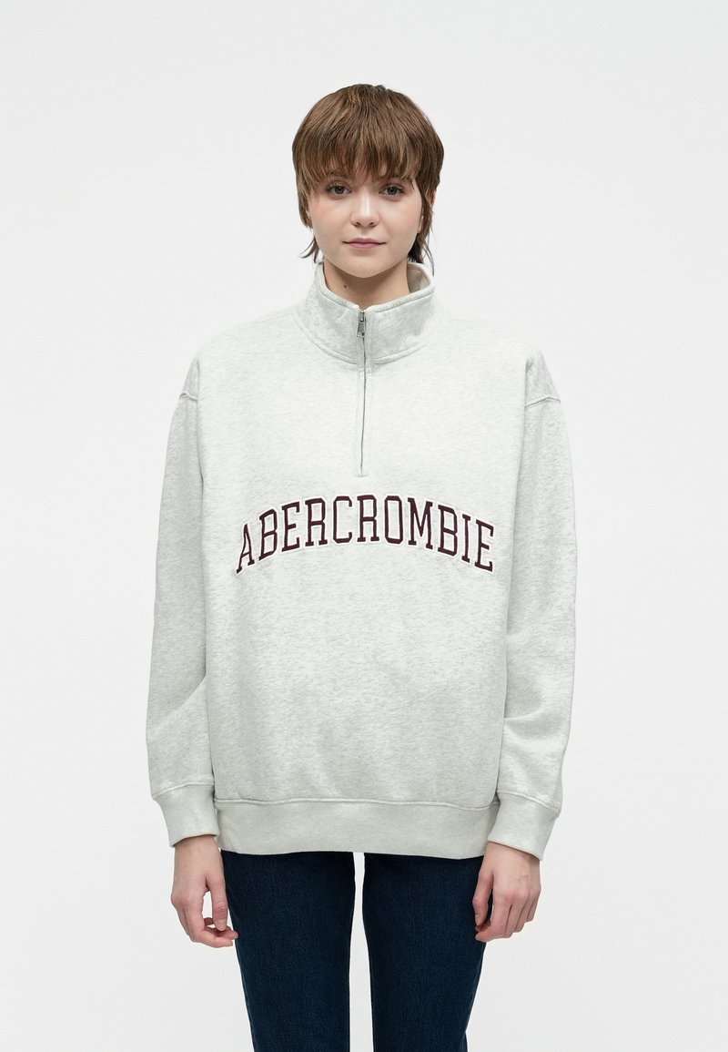 Młoda osoba z krótkimi brązowymi włosami, nosząca jasnoszary sweatshirt z ćwierć suwakowym zamkiem i napisem "ABERCROMBIE" oraz ciemnoniebieskie dżinsy.