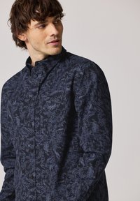 Námořnická košile s knoflíky, zdobená vzorem listů v světlejší barvě. Vyrobena z látky s hladkou strukturou a klasickým límcem.