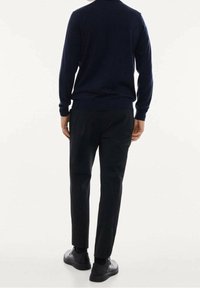 Mann trägt einen marineblauen Pullover, schwarze Hosen und schwarze Schuhe und steht mit dem Rücken zu einem schlichten hellen Hintergrund.