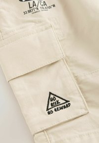 Tissu en toile beige avec une poche appliquée et un logo triangulaire noir brodé contrastant indiquant "PAS DE RISQUE PAS DE RÉCOMPENSE". Texture lisse.