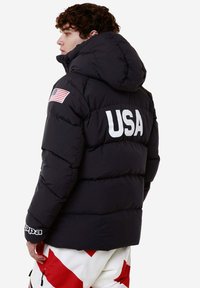 Giacca a vento nera con cappuccio, caratterizzata da un grande testo bianco "USA" sulla parte posteriore e un patch della bandiera americana sulla spalla sinistra.