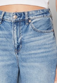 Světle modré džínové kalhoty s vysokým pasem, mají přední kapsy, kontrastní šití a stříbrné zapínání na knoflík. Viditelný detail strukturované tkaniny.