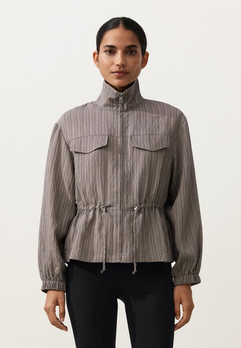 Femme portant une veste grise à fermeture éclair avec col montant, deux poches poitrine, cordons de taille réglables et poignets élastiques, associée à un pantalon noir.