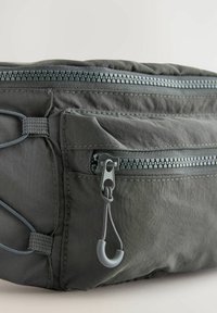 Borsa in tessuto grigio con doppie cerniere superiori, dettagli ad anello e accenti testurizzati. Il materiale sembra resistente e leggermente lucido.