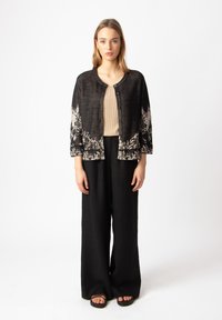 Čierny pletený kardigán s detailným bielym kvetinovým vzorom na leme, skombinovaný s béžovým topom a čiernymi širokými nohavicami. Nosia sa ploché sandále.