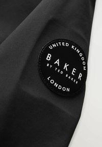 Schwarzer Stoff mit einer strukturierten Oberfläche, auf der ein runder schwarzer Patch mit "BAKER by TED BAKER" in Weiß sowie "UNITED KINGDOM" und "LONDON" zu sehen ist.