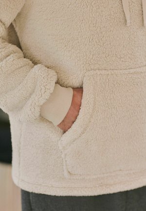 Cream-colored fleece hoodie with a soft, textured surface, featuring a front pocket and ribbed cuffs, partially obscured hand in pocket.