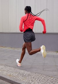 Haut de sport rouge à manches longues avec un motif texturé, associé à un short noir et des chaussures de course blanches avec des accents jaunes et roses.