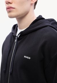 Čierna mikina na zips vyrobená z bavlny s jemnou textúrou, so kapucňou, predným vreckom a bielym logom "HUGO" na hrudi.